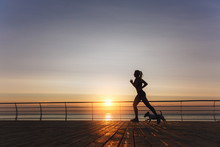 Silhouette Of A Young Beautiful Athletic Girl With Long Blond Hair In Headphones That Runs At Dawn Over The Sea With Her Dog