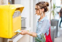 Young Woman Putting Letter To The Old Yellow Mailbox Standing Outdoors On The Street