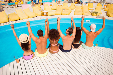 Sticker - Put your hands up! Pool disco party. Six excited cheerful multi ethnic friends are chilling by the pool, sit on the nice wooden white floor, hold arms, luxure resort, yellow beach chairs blue water