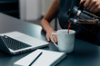 © bnenin - Woman hands pouring coffee into cup on office table.