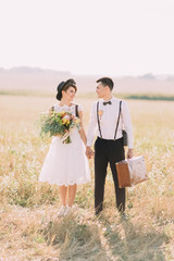 The full-length vertical photo of the vintage dressed newlyweds walking and holding hands in the sunny field. The bride is holding the wedding bouquet and the groom is carrying the old suitcase.