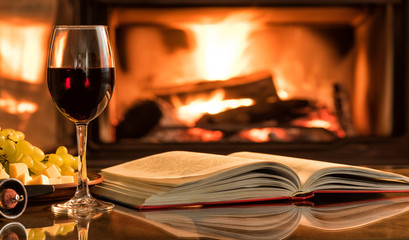 Red wine glass with an open book on table in front of burning fireplace.