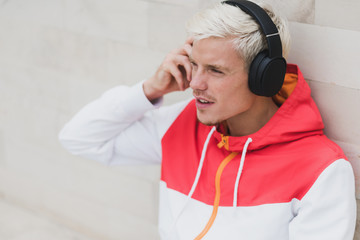 Enjoying attractive portrait of young male dressed in red sportswear resting after physical activities in open air in park.Young handsome man listening to music with headphones against grey background