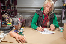 A Young Fair-haired Woman In A Plaid Shirt And Vest Draws A Drawing On A White Pencil Sheet For A Halloween In A Dark Workshop