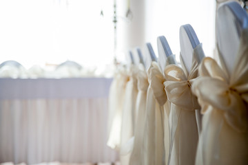 Close-up photo of back of chairs decorated with white cloth cover and big cream satin ribbon tied to a bow
