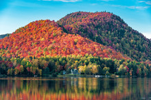 The Hills Covered With Red Maple Forests Behind A Wooden House On The Shore Of A Lake  In Quebec, On A Beautiful Autumn Evening.