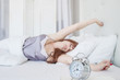 © Надежда Манахова - Beautiful woman stretching in bed. The clock shows 8 o'clock in the morning. White bed and interior. American.