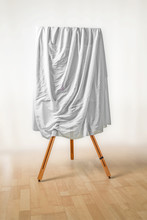 Covered Painting On An Easel, White Cloth Over The Picture, Wooden Floor And Light Background, Art Concept For An Exhibition Opening Day Or A Presentation Ceremony, Copy Space