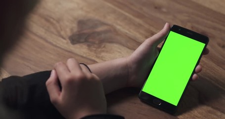 Wall Mural - handheld shot of female teen holding smartphone with green screen over wood table