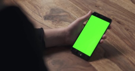 Wall Mural - handheld shot of female teen holding smartphone with green screen over wood table