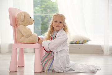 Little girl in medical coat playing with teddy bear and stethoscope on floor