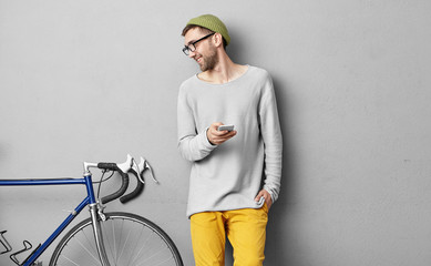 Stylish cheerful young Caucasian hipster with stubble posing indoors and looking at his new bicycle, texting friend on smart phone, boasting about purchase, having happy expression on his face