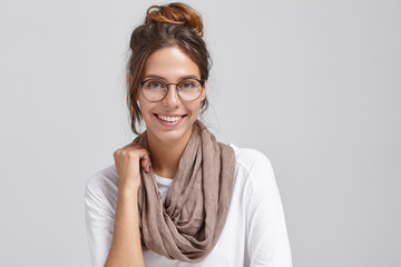 Joyful successful young European female artist or worker of creative profession wearing stylish round glasses and scarf around her neck, looking at camera and grinning broadly, showing white teeth