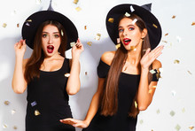 Portrait Of Two Happy Young Women In Black Witch Halloween Costumes On Party Over White Background. Firecrackers In The Background. Confetti . The Concept Of Halloween . Funny Faces
