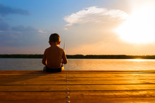 Little Boy Siting On Wooden Dock And Fishing At Sunset.