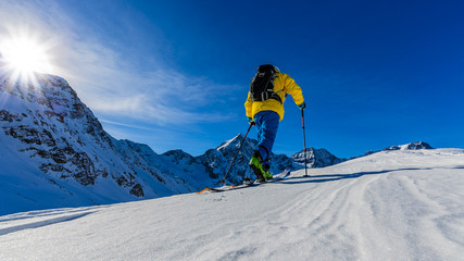 Mountain ski walking up along a snowy ridge with skis in the backpack. In background blue cloudy sky and shiny sun and Tre Cime, Drei Zinnen in South Tirol, Italy. Adventure winter extreme sport.