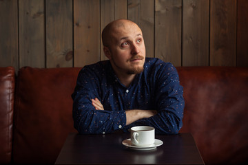 Young serious fashionable man sitting alone in loft-styled cafe. Cup of coffee in his hands. Former factory building, natural daylight.