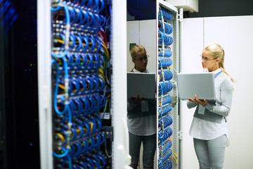 Portrait of young woman working with supercomputer standing in server room holding laptop, copy space