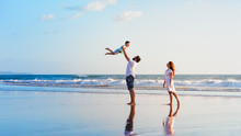 Happy Family Holidays. Joyful Father, Mother, Baby Son Walk With Fun Along Edge Of Sunset Sea Surf On Black Sand Beach. Active Parents And People Outdoor Activity On Summer Vacations With Children.
