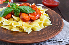 Farfalle Pasta In A Ceramic Bowl, Serve With Ketchup, Fresh Tomatoes, Sausage In Tomato Sauce On A Dark Wooden Background.