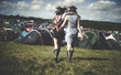 © Mint Images - Rear view of two young women at a summer music festival wearing feather headdresses, walking arm in arm towards tents.