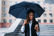 Good news on mobile phone. Moody weather. Happy black female with an umbrella on rainy day, street fashion, happiness concept