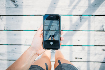 Young social media influencer with marketing ideas makes photo on smartphone of sneaker or running shoes of famous brand, standing on wooden bridge floor