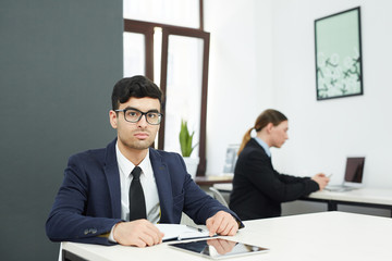 Young serious businessman with notebook and tablet sitting by desk