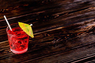 Poster - Top view of glass of red cocktail with ice cubes and straw umbrella on dark wooden table, close-up, copyspace for text