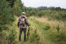 Hunter With A Backpack And A Hunting Gun In The Autumn Forest. The Man Is On The Hunt. Back View.