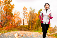 Mature Happy Middle Age Woman Jogging Outside In Her 50s. Middle Aged Asian Chinese Girl In Her Fifties Jogging Outdoor Living Healthy Lifestyle In Beautiful Autumn City Park In Colorful Fall Foliage.