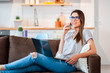 © vladteodor - Casual young woman in a modern decorated home interior sitting comfortable on the couch using phone with laptop.