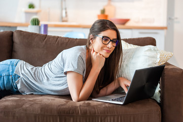 Young creative woman lying on her stomach on the couch using laptop in creative office in a modern decorated home interior