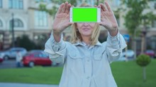 Young Woman Staying In City Park Holds SmartPhone With Pre-keyed Green Screen. Perfect For Screen Compositing. Made From 14bit RAW. 10bit ProRes 444