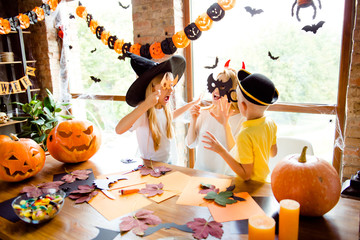 Boo! Blonde mommy with two little dressed sister and brother, creepy costumes, eyewear and head wear, scaring each other in decorated nice loft light room indoors at home near the windows