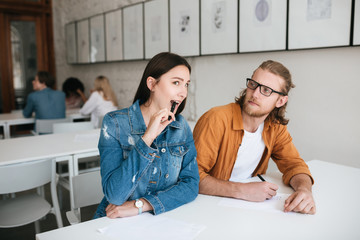 Wall Mural - Pretty girl biting pen while sitting in classroom and trying to write off test in boy that noticed it. Young man sitting at the desk in classroom and thoughtfully looking on girl that sitting near