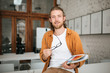 © Anton - Young man sitting on table in office with glasses and document case in hands. Portrait of smiling boy with blond hair and beard joyfully looking in camera while holding documents in hand