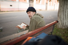 Rear View Of Old Man Sitting On Bench Holding Book. Homeless Man In City Street Reading A Book.