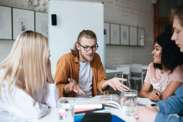 Wall Mural - Portrait of young man with blond hair and beard emotionally explaining something to his friends. Group of people sitting at the table and working together. Students studying with books in classroom