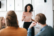 © Anton - Cheerful African American lady with dark curly hair standing near board and happily looking at her colleagues in office. Young beautiful business woman giving presentation to coworkers during meeting