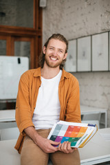 Wall Mural - Portrait of young man sitting on desk in auditorium with document case in hands. Cheerful boy with blond hair and beard  happily looking aside while holding journals in hands