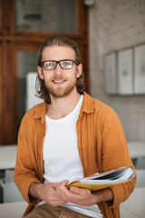 Portrait of young smiling man sitting on desk in auditorium with document case in hands. Cheerful boy with blond hair and beard in glasses happily looking in camera while holding journals in hands