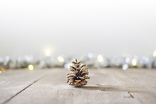 Pine Cone Sitting On Rustic Wooden Surface With Holiday Lights And Tinsel In Soft Focus