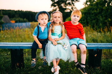 Wall Mural - Group of little emotional kids sitting on bench outdoor in countryside. Girl in dress between two guys. Difficult relations. Youth jealousy. Funny loving triangle. Joy, sorrow, hurt and offence.