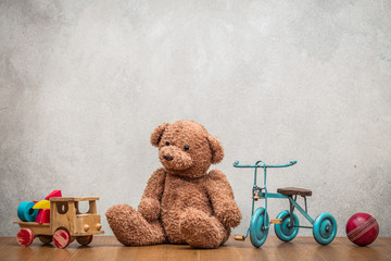 Retro plush Teddy Bear, old toy trike bicycle, obsolete wooden truck with construction blocks and leather ball front concrete textured wall background. Vintage style filtered photo