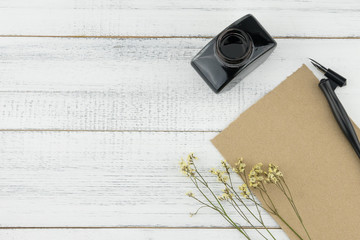 Blank brown card, oblique pen and bottle of ink decorated with yellow limonium caspia flowers on white wood background 