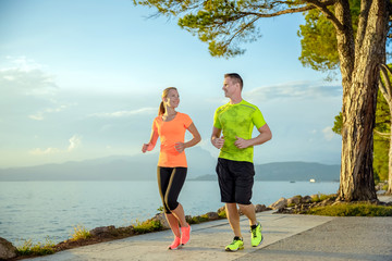 Young sexy couple is running along the promenade. they are doing their sport workout in the beautiful sundown along the beach. colorful dress, trees, water, mountains and a amazing blue sky.