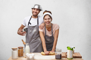 Wall Mural - Happy friendly family bakers at kitchen together: smiling beautiful housewife kneads pastry for cake and attractive funny man stands near his wife, being involved in culinary process. Busy cookers