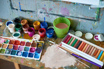 Background image of messy art supplies set on shelf of artists easel: watercolor and gouache paints,  pastel chalk next to used paintbrushes and cup of water