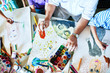 © Seventyfour - Top view of childrens drawings on table in art class with group of people painting and teachers hands